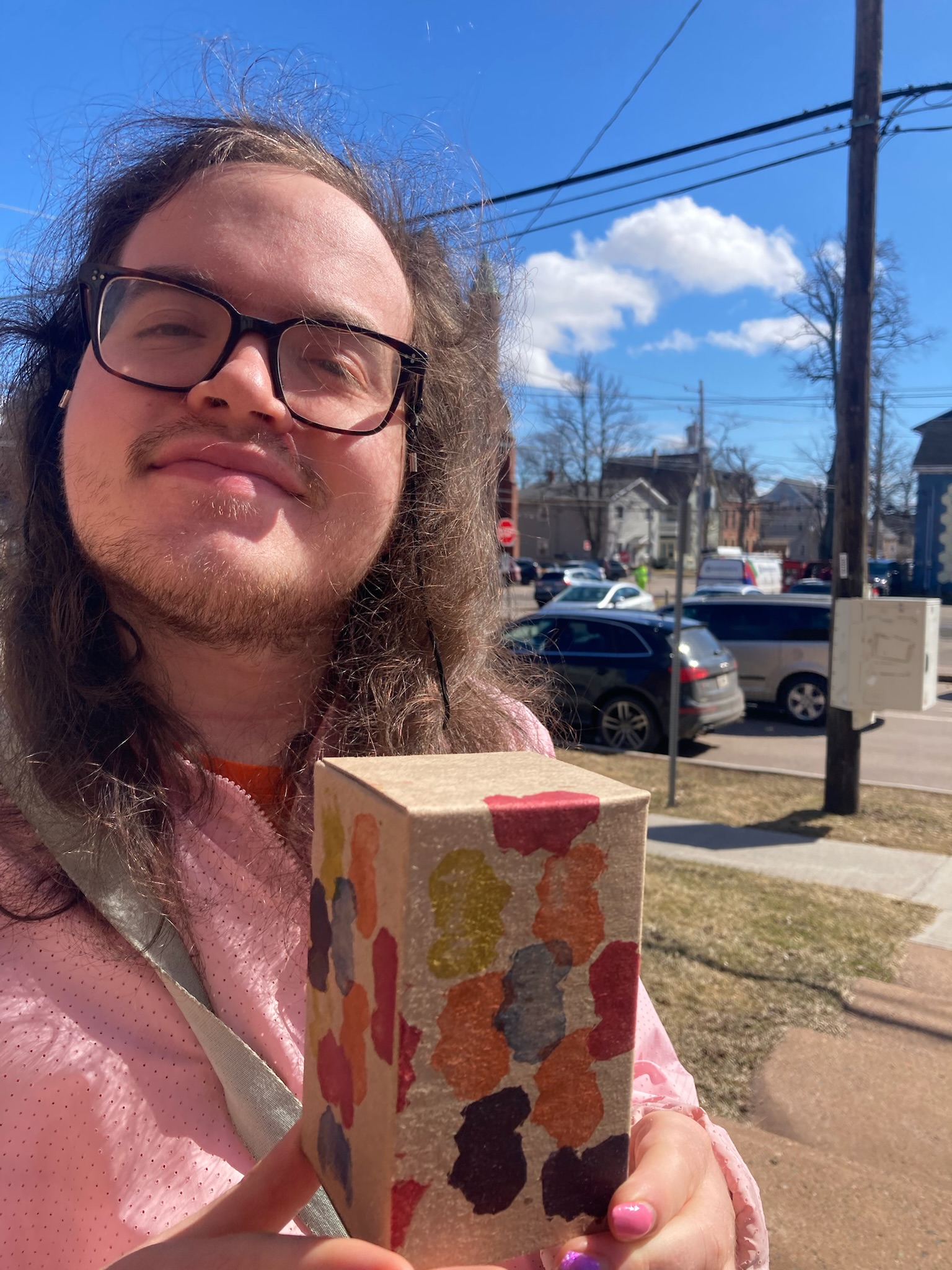 Olivia holding the prototype box in her hand, standing outside on the sidewalk on a bright spring day, wearing a pink windbreaker, with a smile on her face.