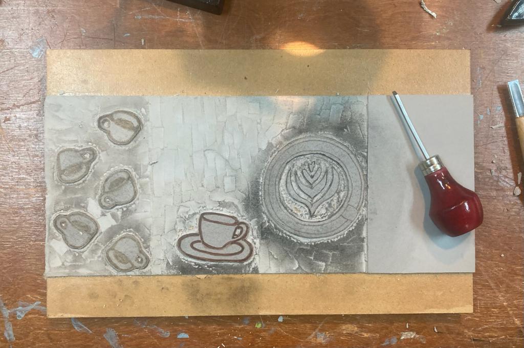 A 12 inch by 5 inch piece of mounted linoleum block divided into four sections, with three of the four sections carved with coffee scenes, and the final panel, on the right, still to be carved.