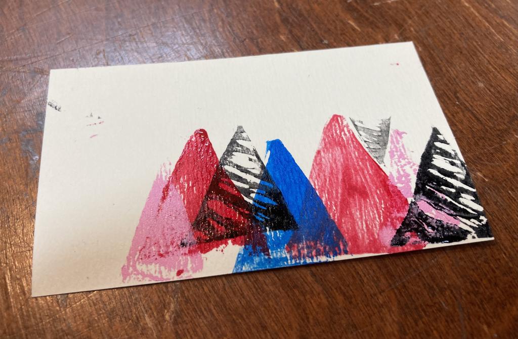 Simple trees-as-triangles, rendered in block printing on a piece of card, sitting on a table.