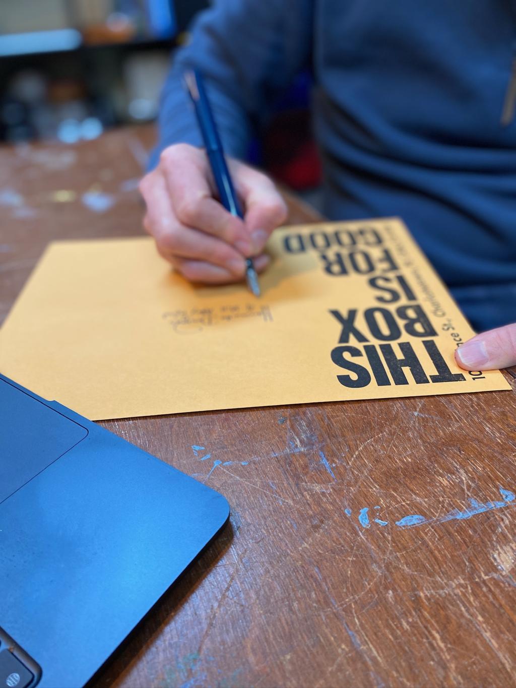 A blurry photo of me writing an address on a THIS BOX IS FOR GOOD-printed envelope, on a brown wooden table.