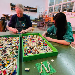 Two people, sitting at a table in front of large boxed of Lego