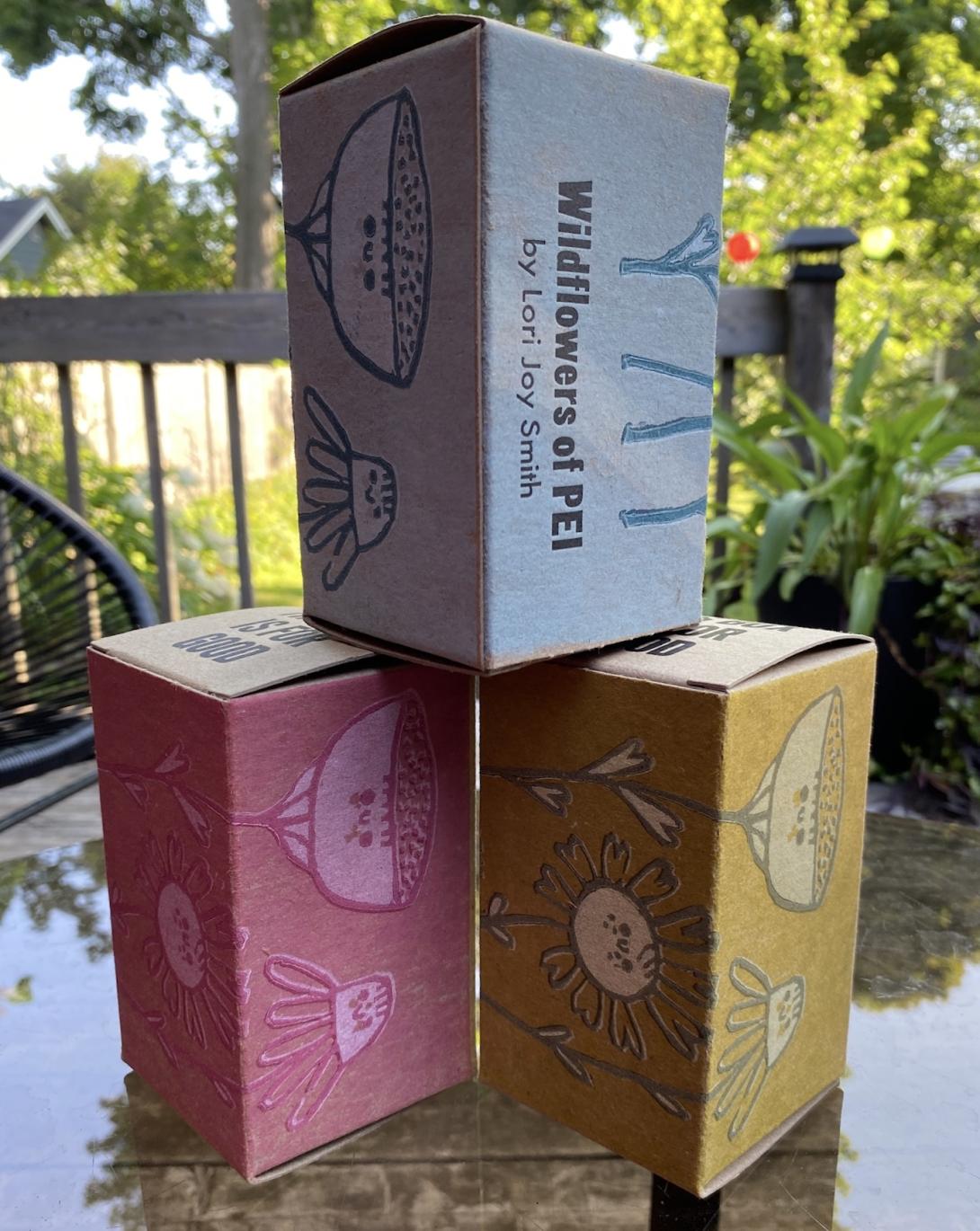 Three of our August boxes, stacked one-on-two, on a glass tabletop. The bottom two boxes are pink and yellow; the top box is blue. They each show a different side of the box, with stylized renderings of PEI wildflowers.